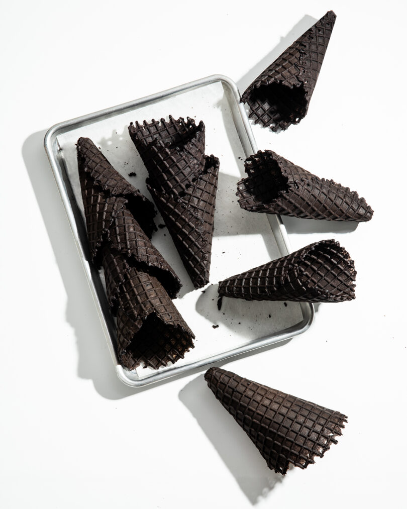 A metal tray and white surface displaying eight empty dark chocolate waffle cones, some on the tray and some scattered around it.