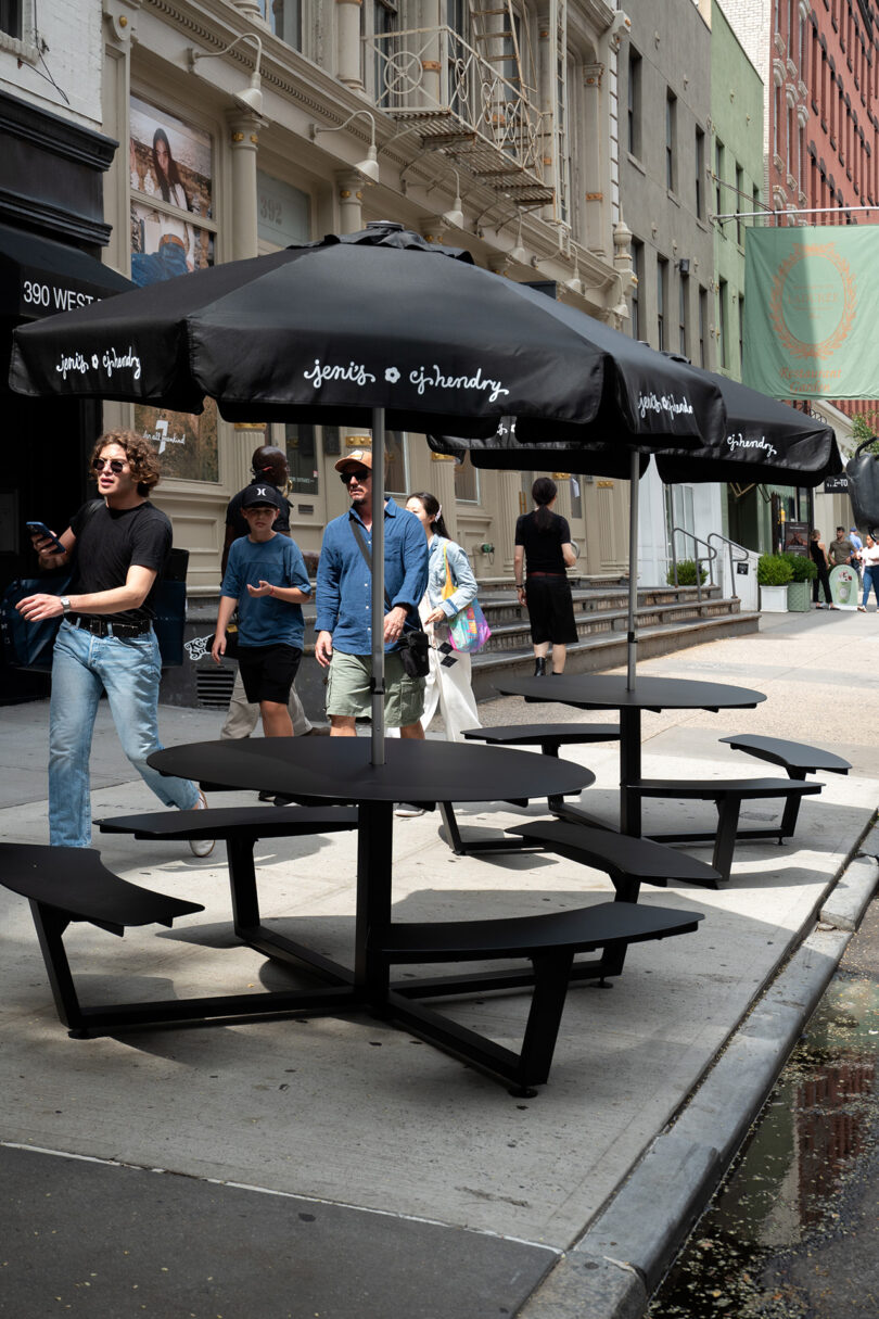 A group of people walk past empty black picnic tables with umbrellas from the OPAQUE CJ Hendry collection on a city sidewalk next to storefronts.
