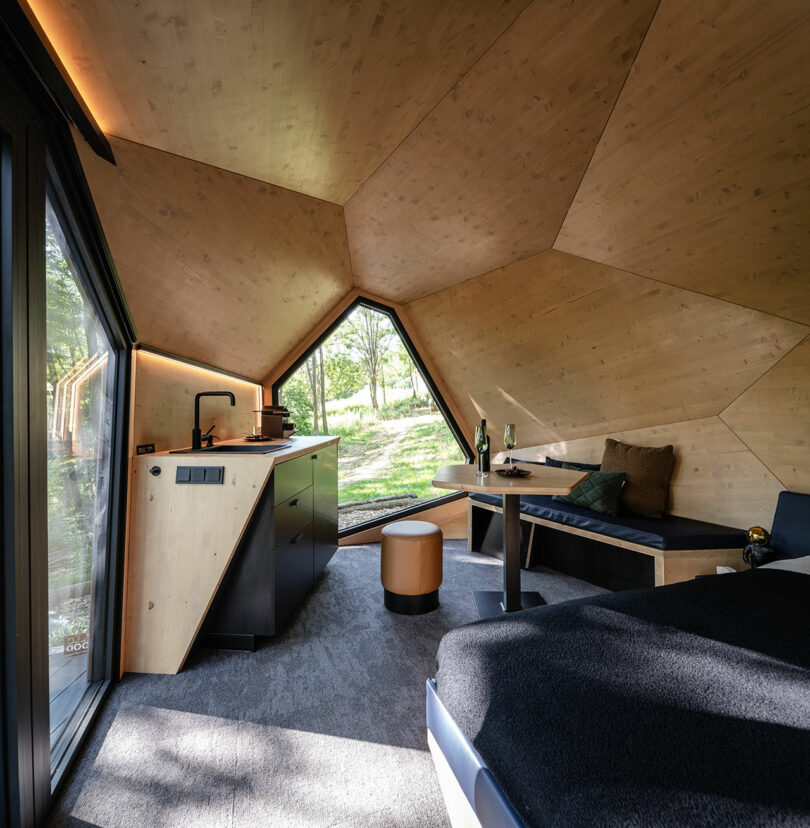 Un interior moderno de cabina con paneles de madera con un techo geométrico, cocina, asiento de banco y una gran ventana triangular con vistas a la vegetación.