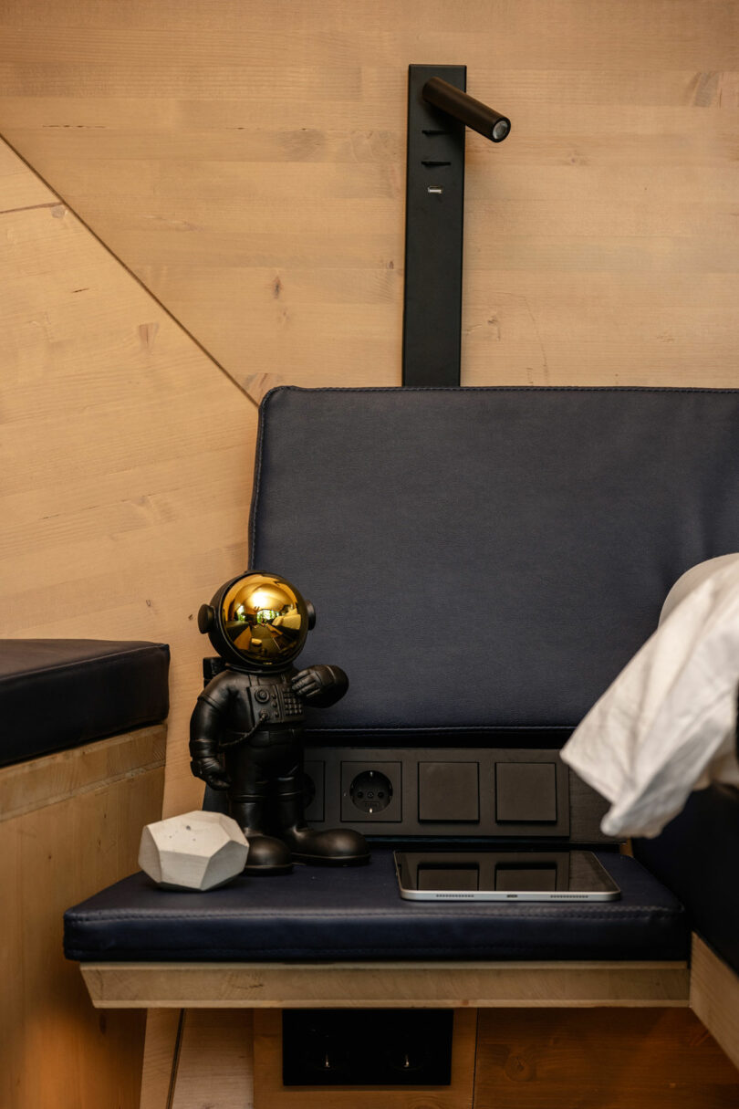 Una pequeña figura de astronauta con un casco de oro se encuentra en un banco al lado de un objeto blanco, salidas de energía y una cama con cojines azules en una habitación con paneles de madera.