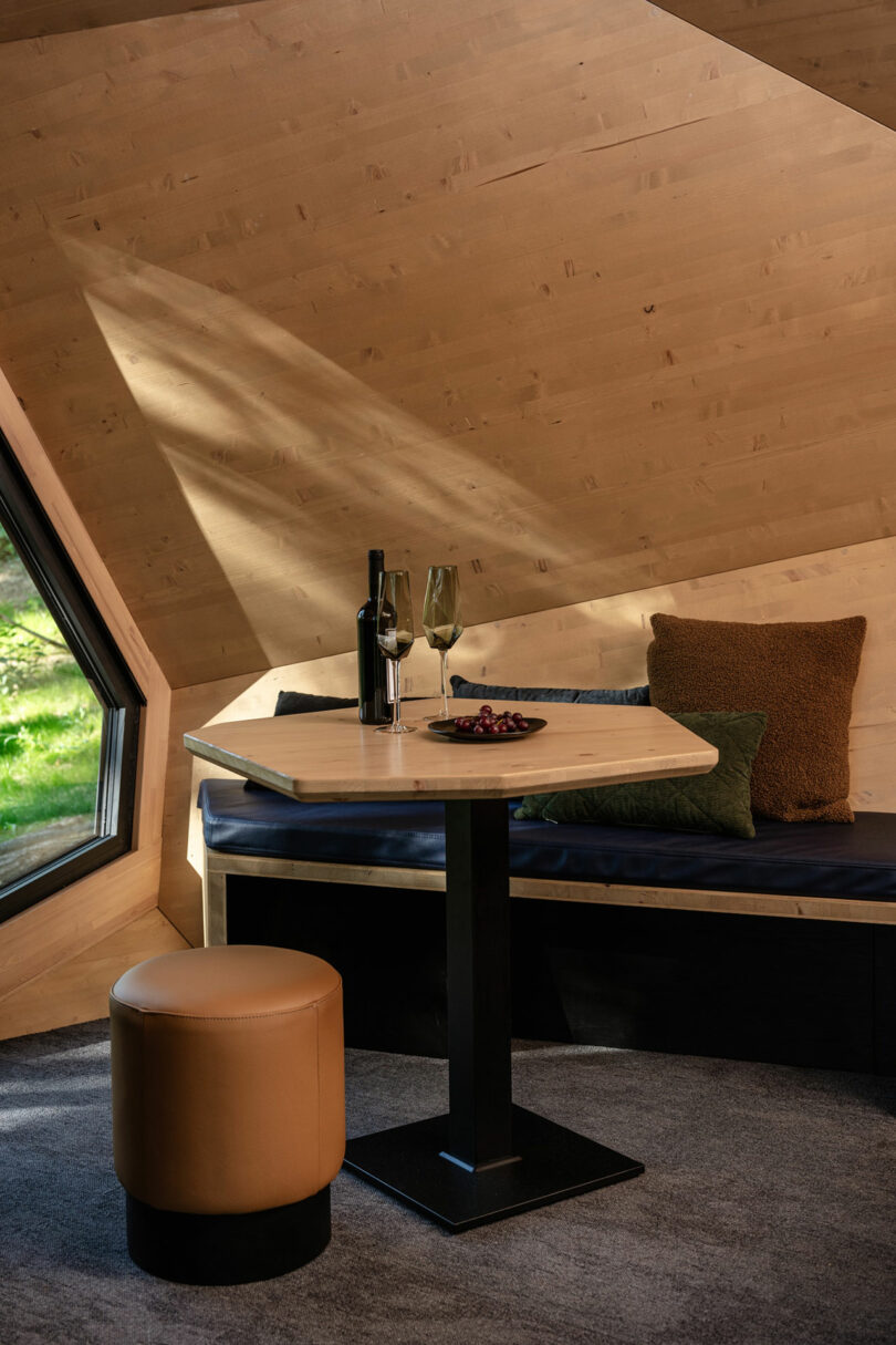 Una pequeña mesa de madera con una botella de vino, dos copas y un plato de uvas se encuentra al lado de un banco con almohadas y un taburete marrón redondo en una acogedora habitación con paneles de madera.
