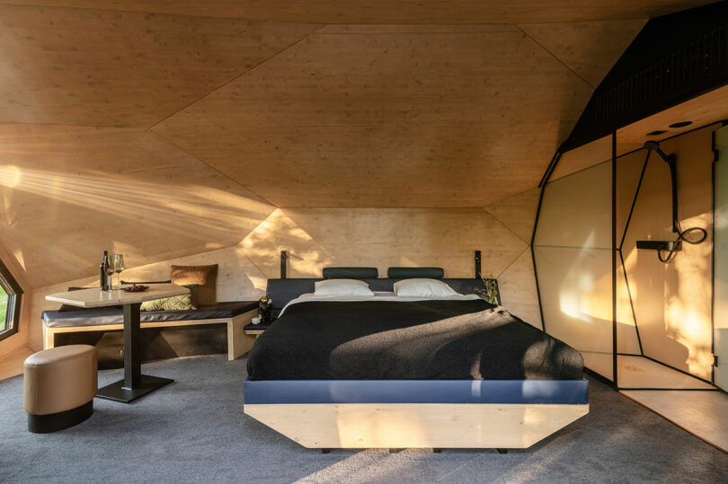 Dormitorio moderno con paredes y techo con paneles de madera, una cama grande con ropa de cama oscura, una pequeña mesa de comedor y una área minimalista de ducha abierta. La luz natural entra desde una ventana.