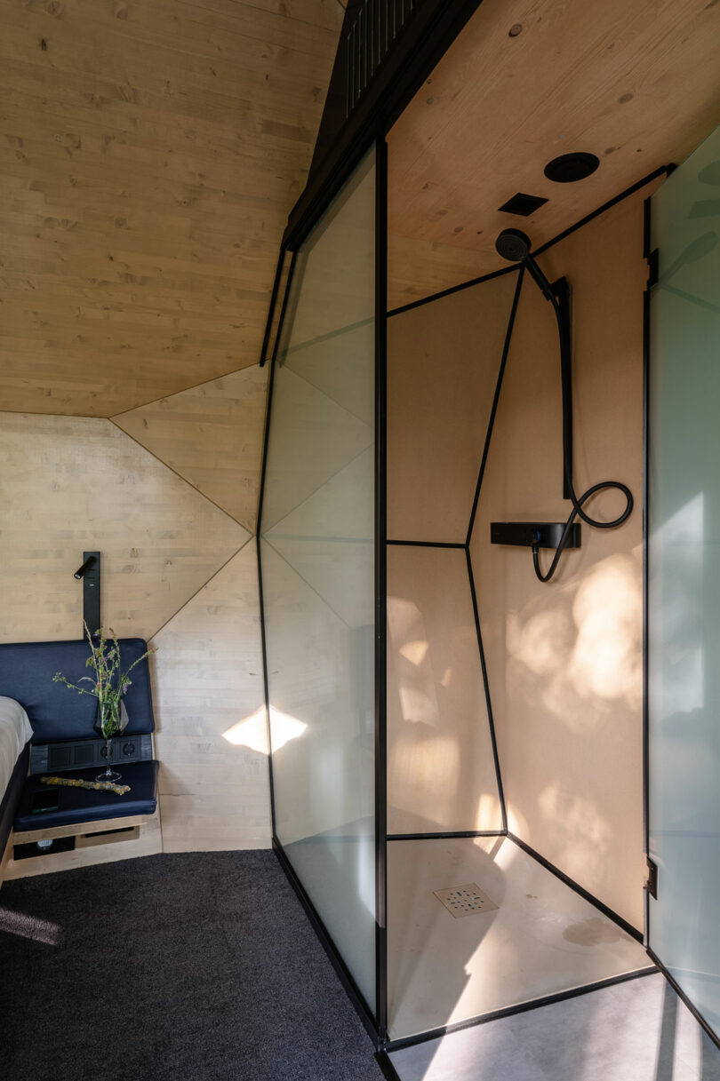 Baño minimalista con una ducha engrasadas de vidrio, accesorios negros, paredes de madera clara y un pequeño estante de noche que sostiene un jarrón de vidrio con vegetación.