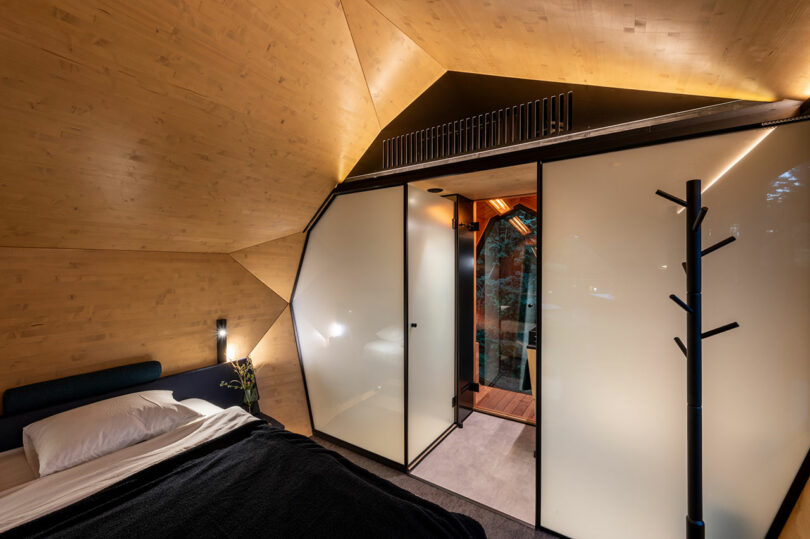 Dormitorio moderno con un techo de madera, partición de vidrio esmerilado, decoración minimalista y una cama visible con ropa de cama blanca y una manta oscura.