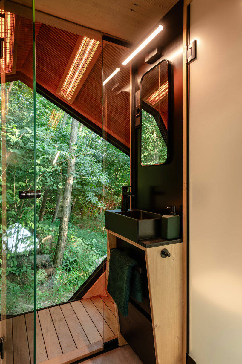 Un baño moderno con un fregadero negro, acentos de madera, paredes de vidrio y una gran ventana con vista a un exuberante bosque.