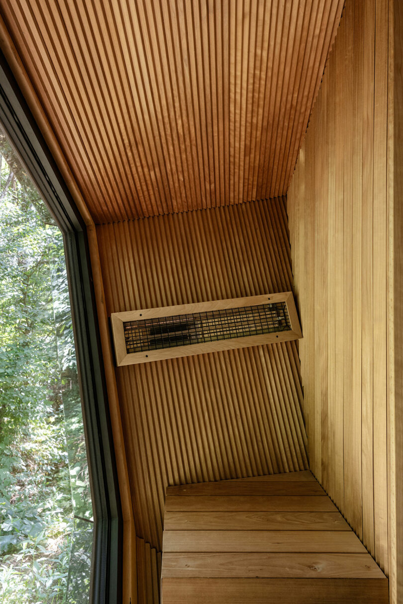 Muro y techo interior con paneles de madera con una ventilación del calentador, al lado de una gran ventana con vistas a los árboles verdes afuera.