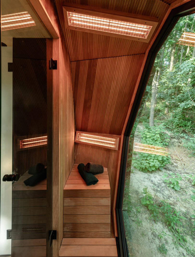 Una moderna sauna de madera con calentadores incorporados, un banco y dos toallas enrolladas, con una gran ventana con vistas a un área boscosa.