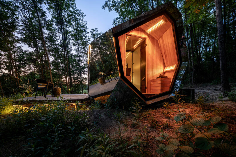 Una cabaña pequeña y moderna con diseño angular e interior de madera iluminado se encuentra en un área boscosa al anochecer, rodeada de árboles y muebles de exterior.