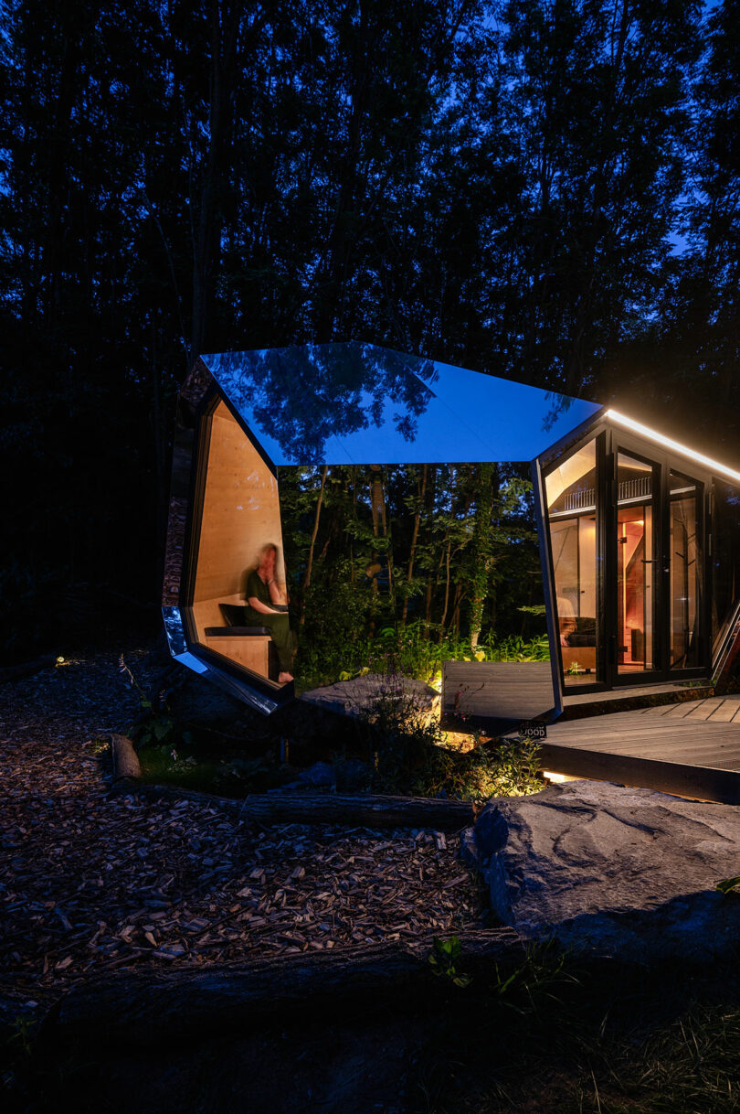 Una cabaña de vidrio moderna y angular con interior iluminado se encuentra en un área boscosa al anochecer; Una persona es visible por dentro, sentada cerca de una ventana.