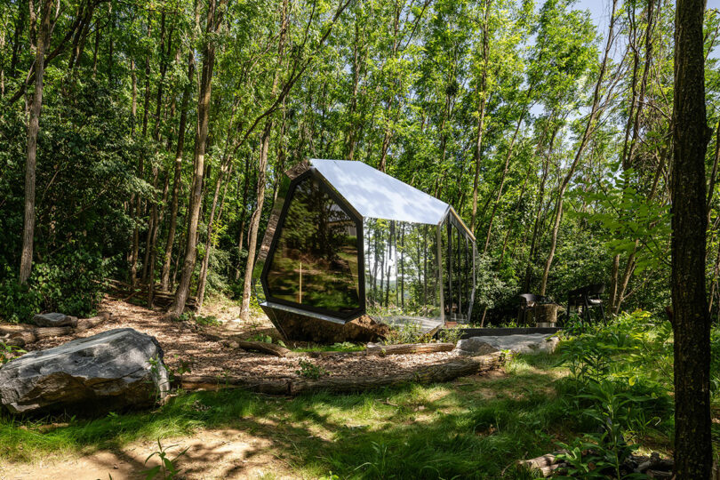 Una pequeña cabina moderna con paredes de vidrio reflectantes y una forma geométrica se encuentra en una zona boscosa rodeada de árboles altos y grandes rocas.
