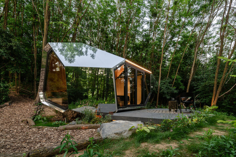 Una cabaña de vidrio moderna y angular con superficies espejadas se encuentra entre los árboles en un bosque, con un patio con una parrilla en una cubierta de madera.
