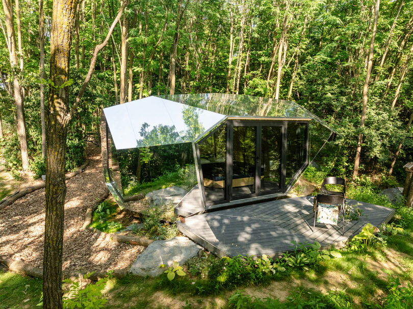 Cabina moderna con paredes exteriores espejadas y grandes puertas de vidrio, situada en una cubierta de madera en un exuberante y verde limpieza.
