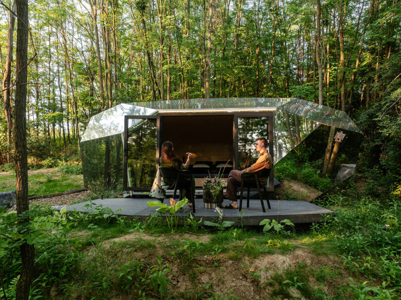 Dos personas se sientan y hablan en una pequeña cubierta de madera fuera de una cabaña moderna con paneles de vidrio rodeada de denso bosque verde.