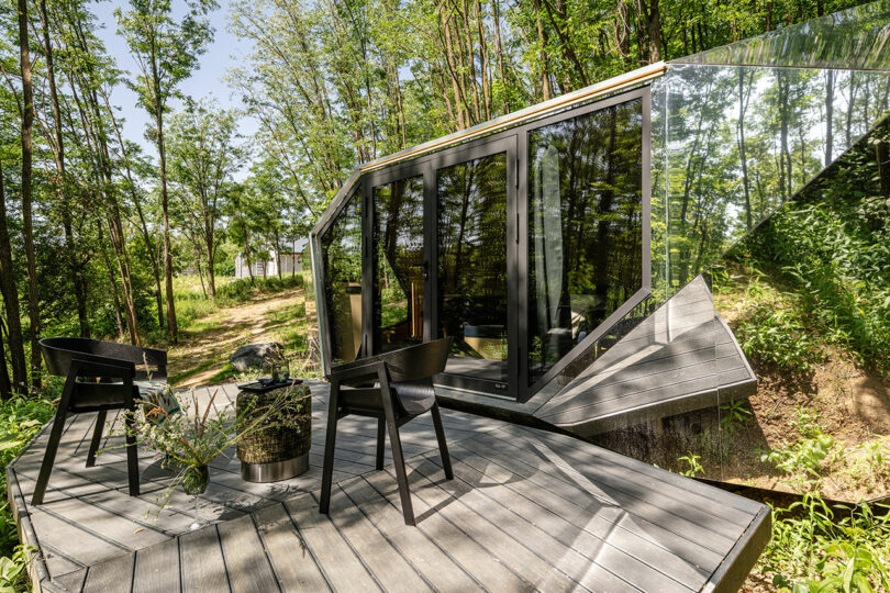 Cabina moderna con grandes ventanas de vidrio y diseño angular, ubicado en un área boscosa, con una pequeña cubierta con dos sillas y una mesa.