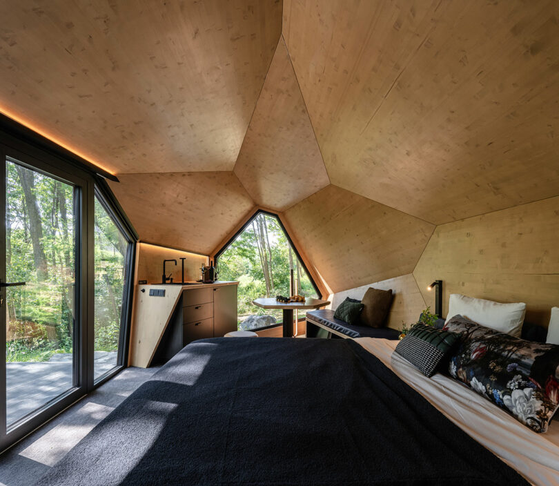 Dormitorio moderno con techo de madera geométrica, grandes ventanas con vistas a los árboles, una cama con ropa de cama oscura, una pequeña área de cocina y una mesa con dos sillas. La luz natural llena el espacio.