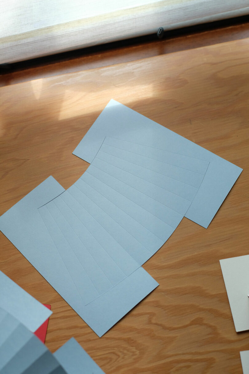 A piece of white paper with evenly spaced lines and two rectangular extensions lies on a wooden surface near a window.
