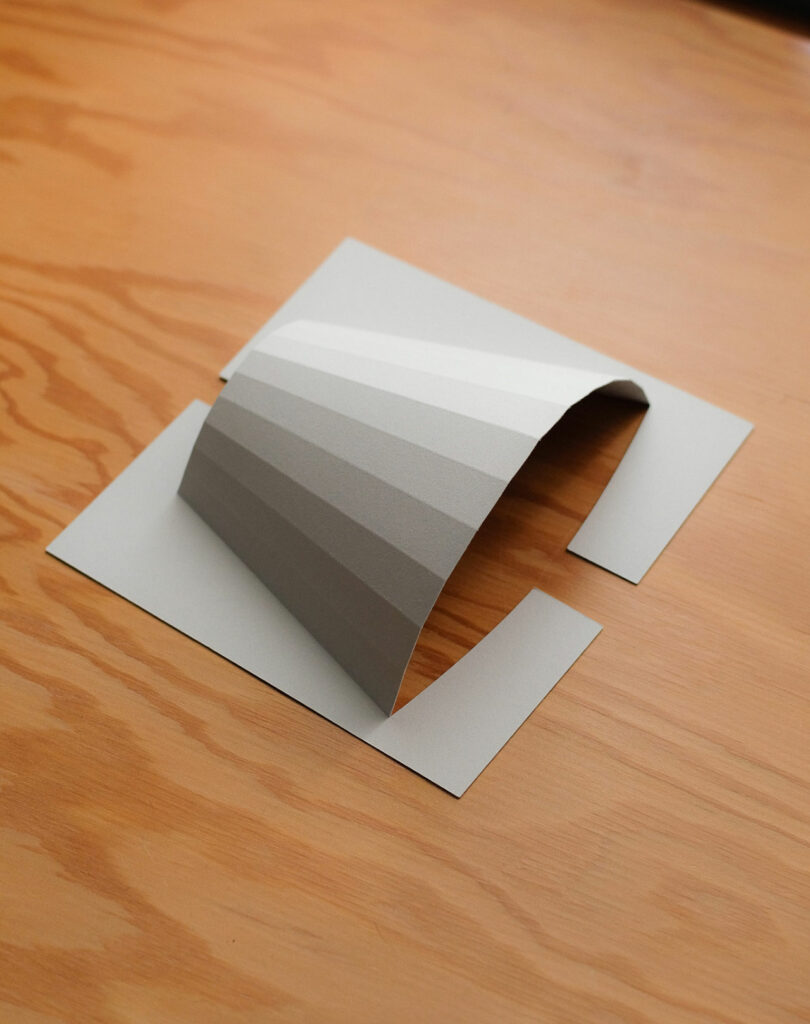 A piece of gray paper is folded in an arch shape, resting on a wooden surface with two rectangular cutouts beneath each end of the arch.