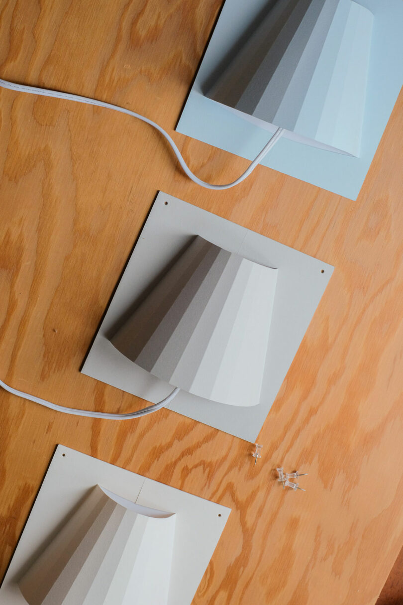 Three wall-mounted lamps with gradient gray shades are displayed on a wooden surface, each with a white cord and screws placed nearby.