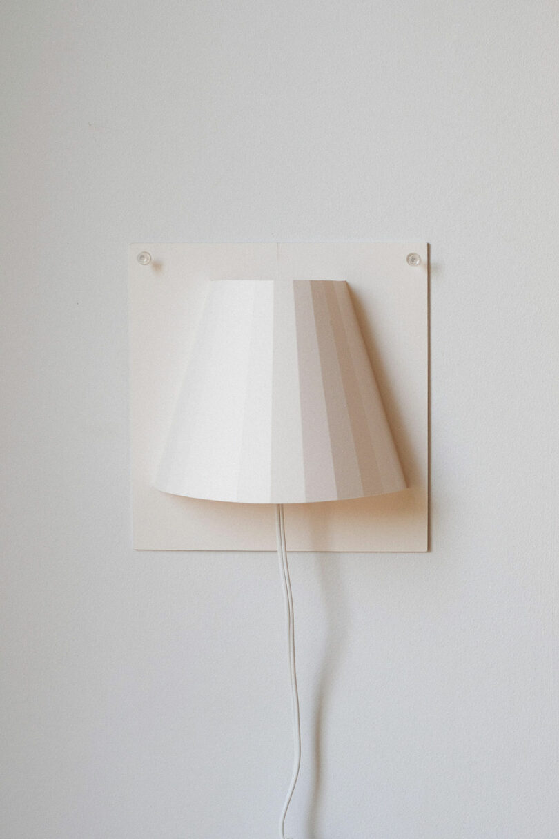 A white wall-mounted lamp with a pleated shade and a visible cord hanging down, positioned against a plain light-colored wall.