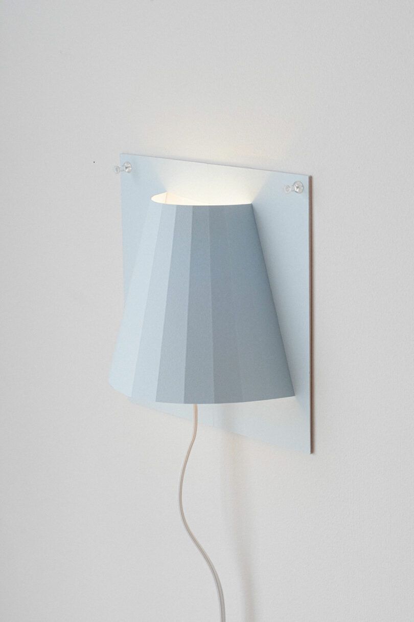 A modern wall-mounted lamp with a white, angular lampshade, attached to a rectangular wall plate, and a visible white power cord hanging down.