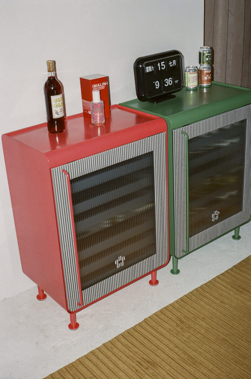 Dos gabinetes de metal, uno rojo y uno verde, se paran uno al lado del otro en la pared. El gabinete rojo tiene una botella y una caja en la parte superior, y el gabinete verde tiene productos enlatados y un reloj digital en la parte superior.