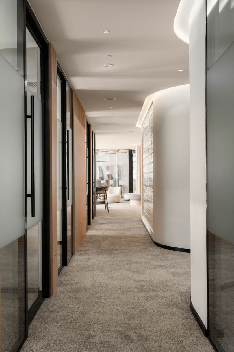 Modern office corridor with glass wall with the left, curved wall on the right, carpet floor and seating, visible at the end near large windows.
