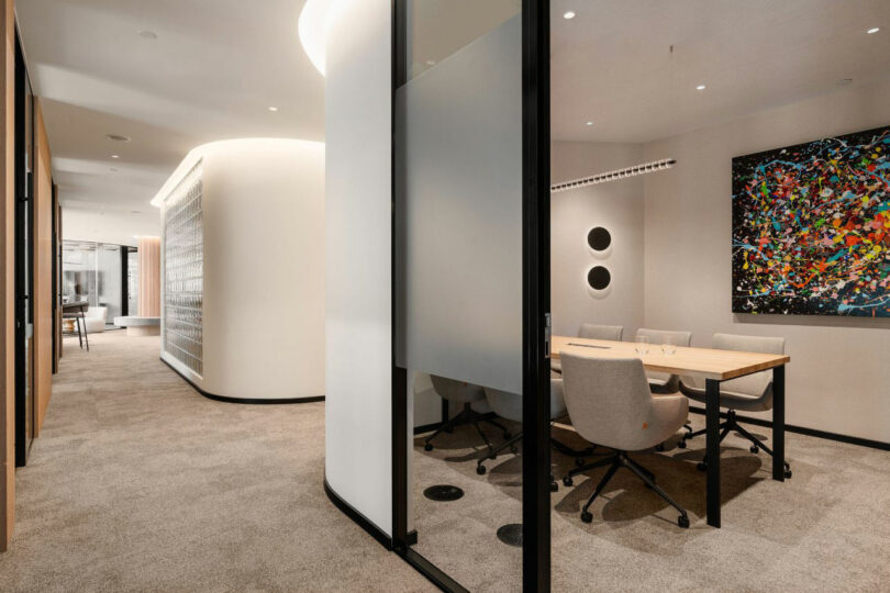 Contemporary office with them with a glass hallway, four chairs around the table, abstract art on the wall and carpet in a neutral color scheme.