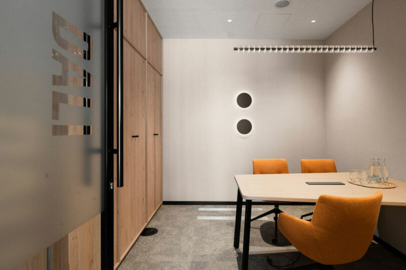 Mala modern meeting room with wooden table, two orange chairs, wall shelves, round lyrics on wall and glass door marked "Dali." Three bottles with glass waters are sitting on the table.