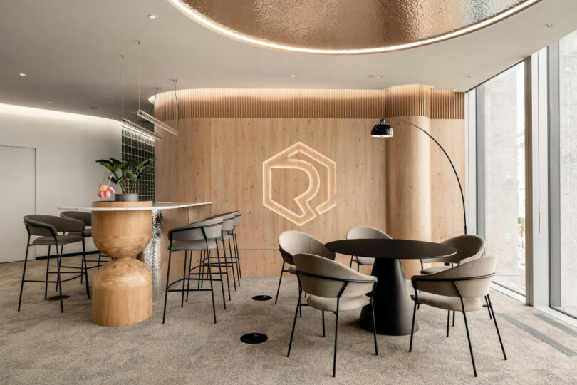 Contemporary office with round table, chairs, high chairs on the bar, and a wooden wall containing a geometric logo. Large windows provide natural light.