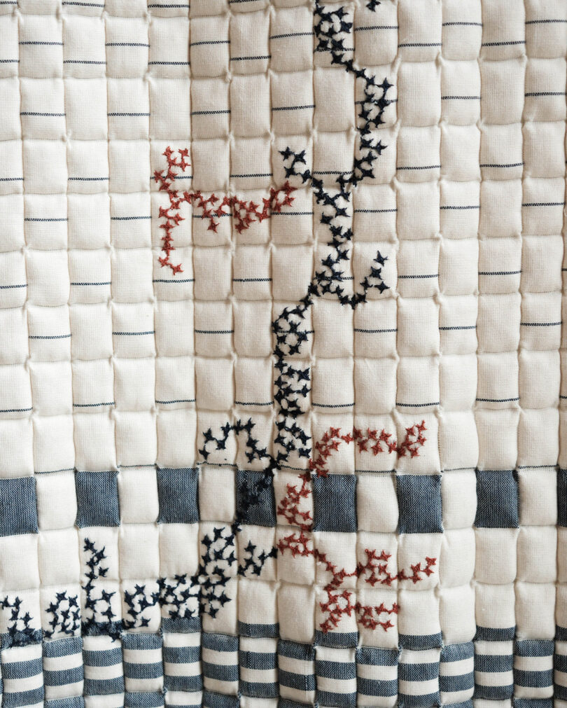 Un textil acolchado con costuras verticales y horizontales decorado con patrones de punto de cruz en negro y rojo formando formas abstractas.