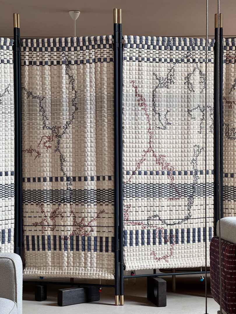 Divisor de habitación de tela con patrón de tablero de ajedrez tejido en blanco y azul, sostenido por varillas verticales negras, ubicado entre muebles modernos en un interior moderno.