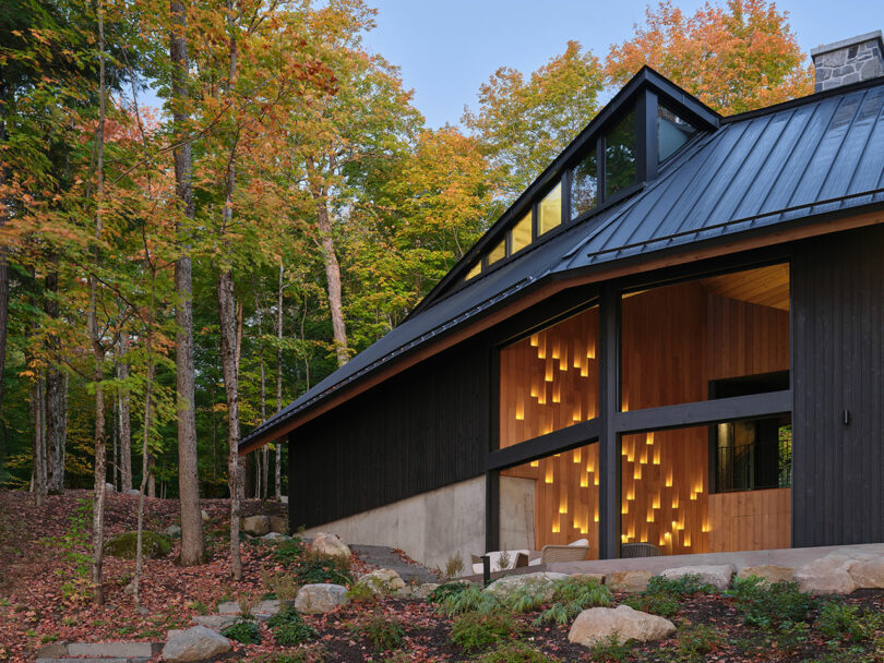 Modern house with black siding and large windows, set among trees with autumn foliage; warm lights illuminate part of the wooden facade.