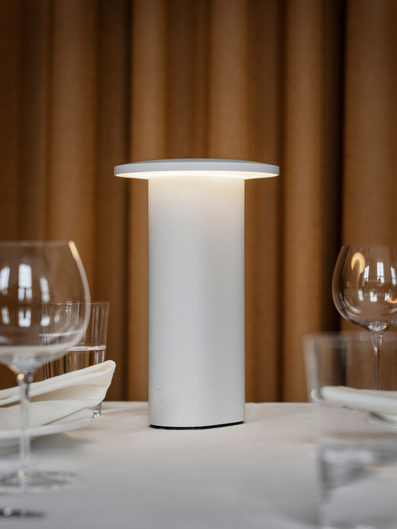 Una lámpara de mesa cilíndrica blanca que recuerda a los diseños minimalistas de Cecile Manz arroja un suave brillo sobre una mesa de comedor amueblada con cristalería, platos y servilletas dobladas. Las cortinas marrones crean un telón de fondo cálido.