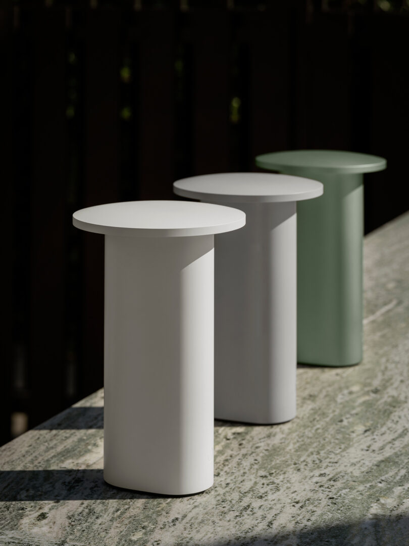 Tres mesas auxiliares cilíndricas de Cecile Manz, rematadas con círculos planos en blanco, gris y verde, se alinean en fila sobre una superficie de mármol al aire libre.