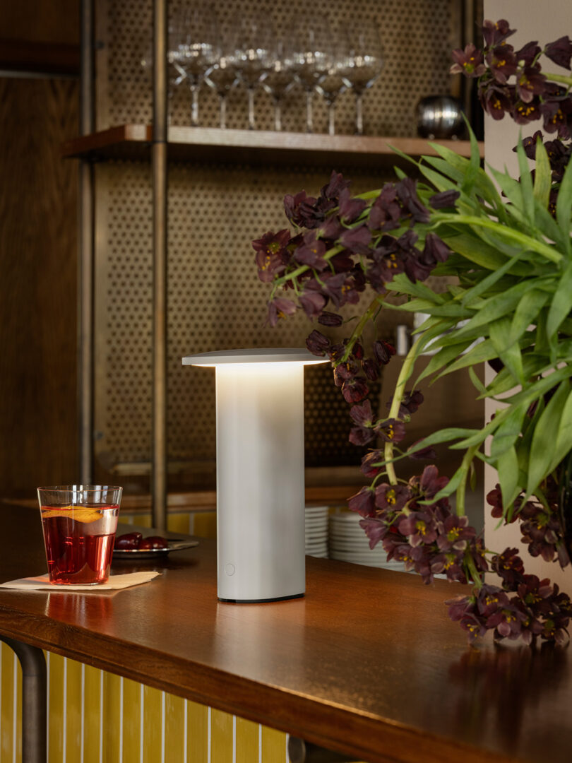 Una moderna lámpara de mesa blanca de Cecile Manz se encuentra sobre un mostrador de madera junto a una bebida roja sobre un fondo de flores y cristalería de color morado oscuro.