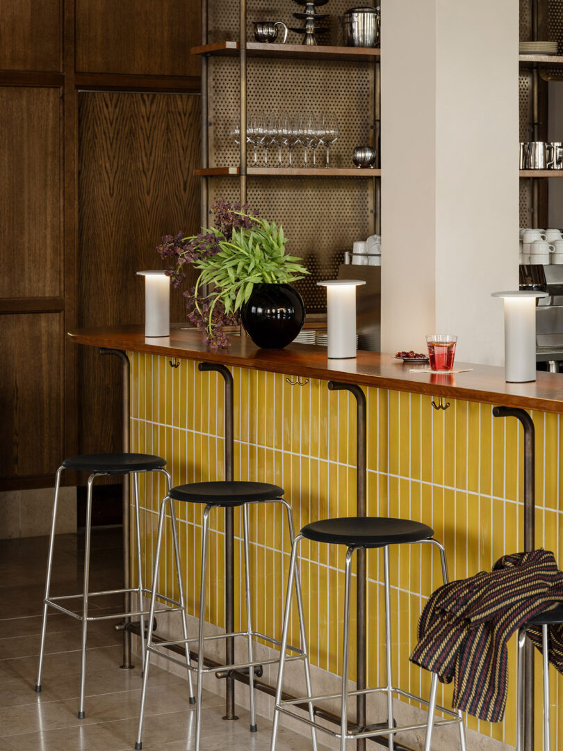El frente de la barra está pavimentado con azulejos amarillos, tres taburetes negros de Cecile Manz, un taburete cubierto con tela a rayas y la encimera de madera está cuidadosamente dispuesta con plantas, vasos y bebidas.