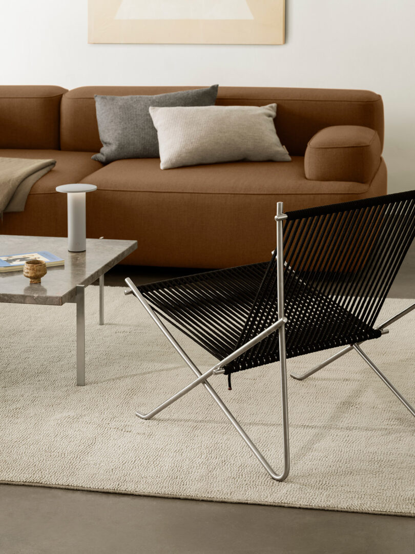 Sala de estar moderna con sofá marrón, cojines grises y beige, mesa de centro de mármol y una silla de hilo negro inspirada en Cecile Manz apoyada sobre una alfombra de textura clara.