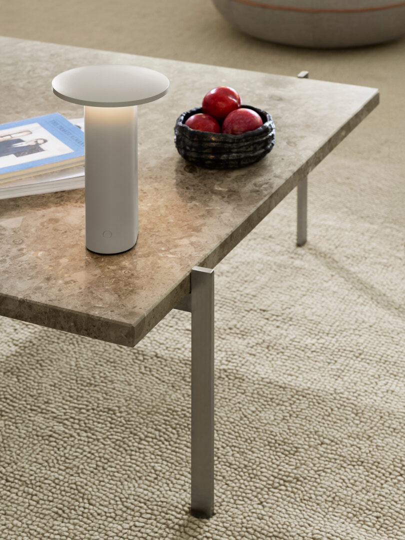 Una mesa de centro de mármol con una moderna lámpara blanca de Cecile Manz, un cuenco de manzanas rojas negras y una revista descansa sobre una alfombra beige texturizada.