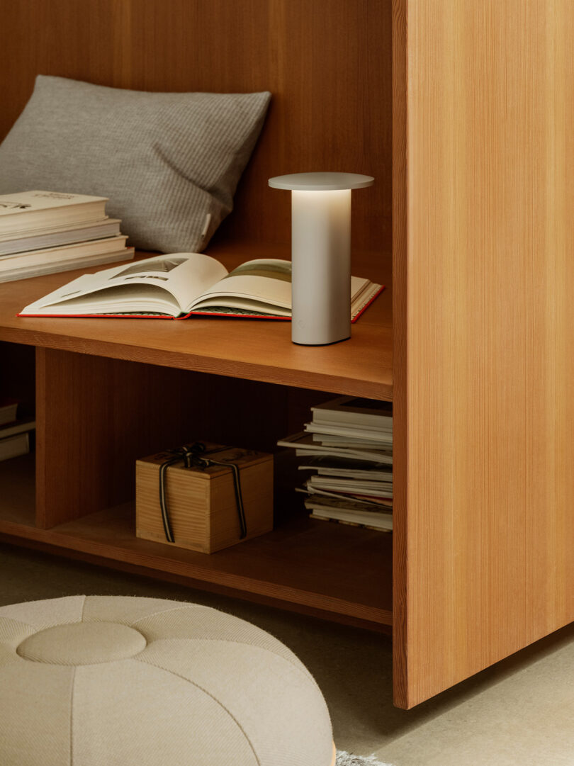 Una lámpara de mesa moderna de Cecile Manz se encuentra junto a un libro abierto en un estante de madera, con un cojín, libros apilados y una caja de madera cerca.