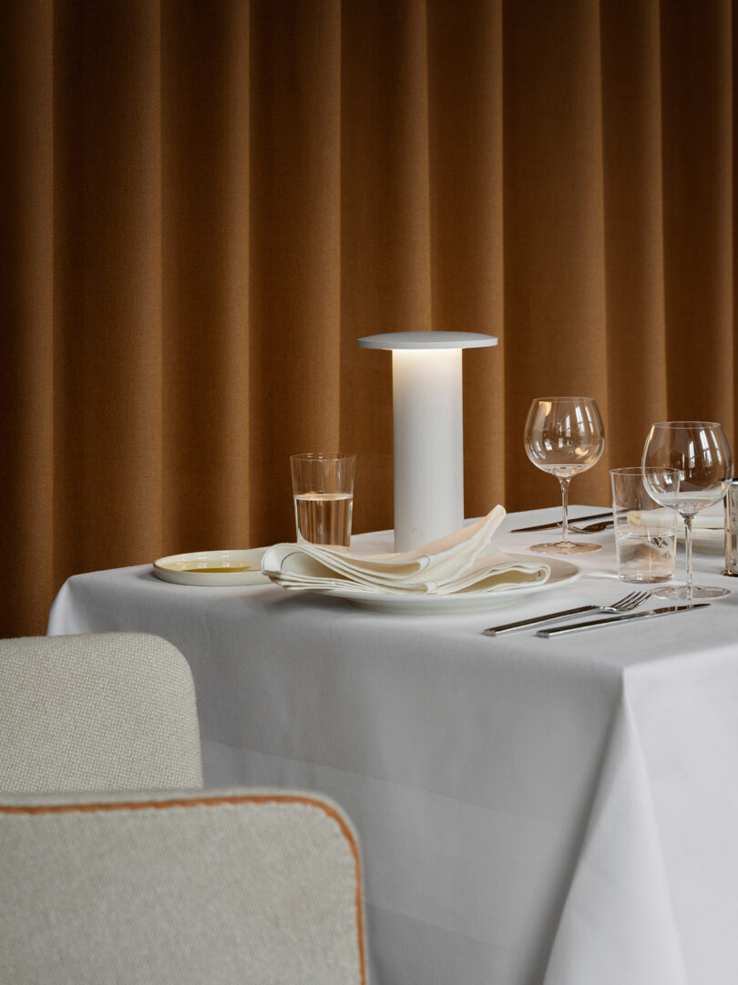 La mesa está cuidadosamente dispuesta con servilletas dobladas, cristalería, cubiertos y una moderna lámpara blanca de Cecile Manz combinada con cortinas marrones.