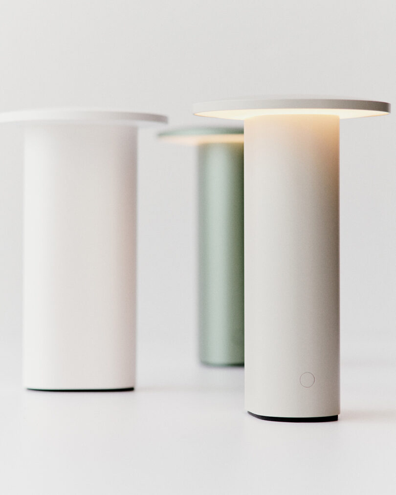 Tres sencillas lámparas de mesa cilíndricas planas, blancas y verdes, diseñadas por Cecile Manz, se exponen sobre una superficie blanca, una de las cuales está iluminada.