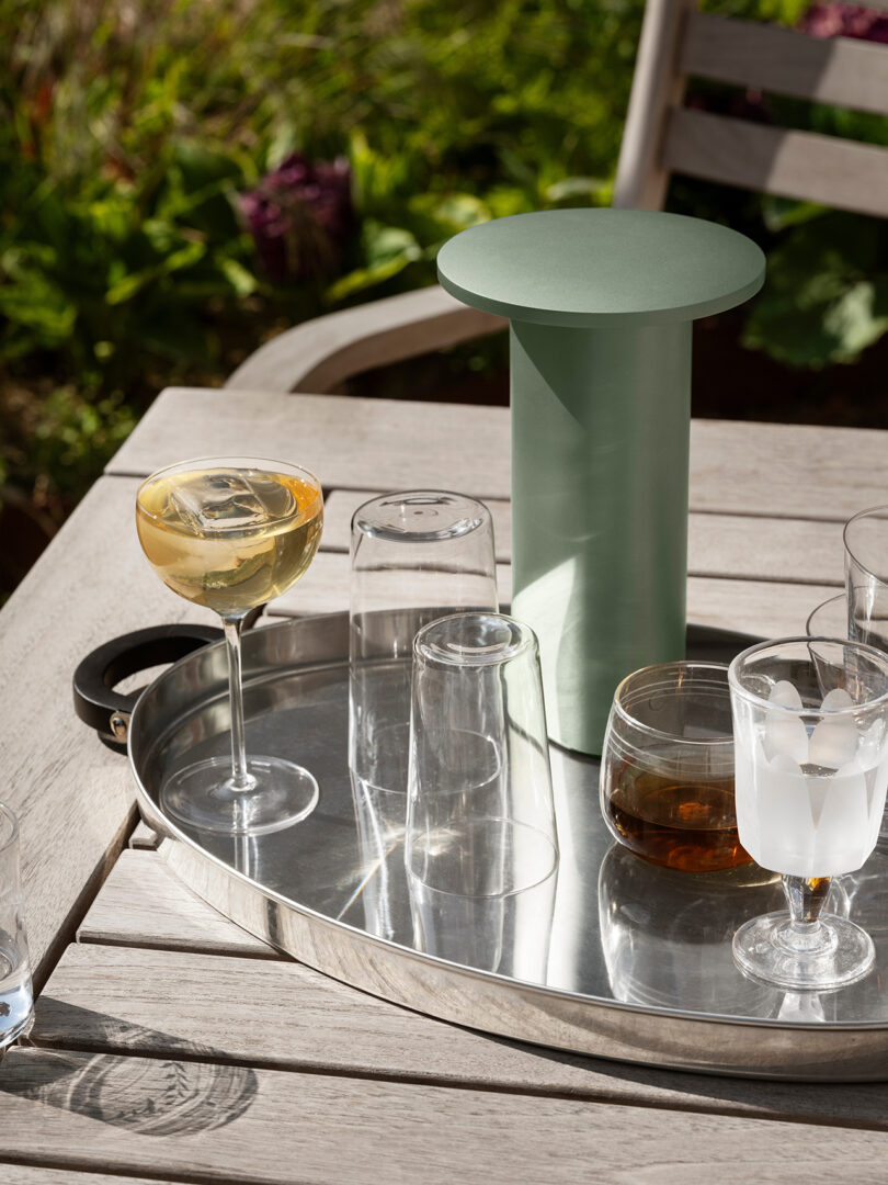 Una bandeja de metal se encuentra sobre una mesa de madera al aire libre, con vasos, un cóctel y un objeto cilíndrico verde diseñado por Cecile Manz, rodeado de un exuberante follaje.