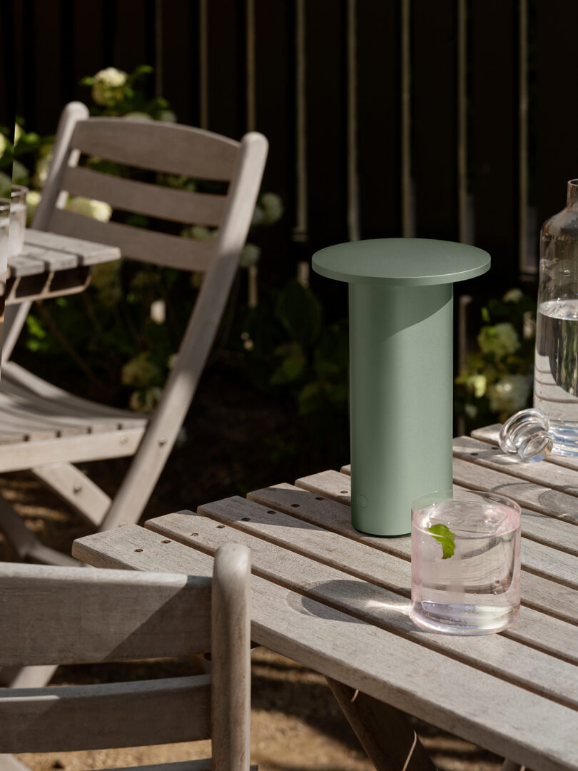 Un objeto cilíndrico de color verde mate, que recuerda a los diseños minimalistas de Cecile Manz, se encuentra sobre una mesa de madera al aire libre junto a un vaso de agua y una hoja, rodeado de sillas de madera y exuberante vegetación.