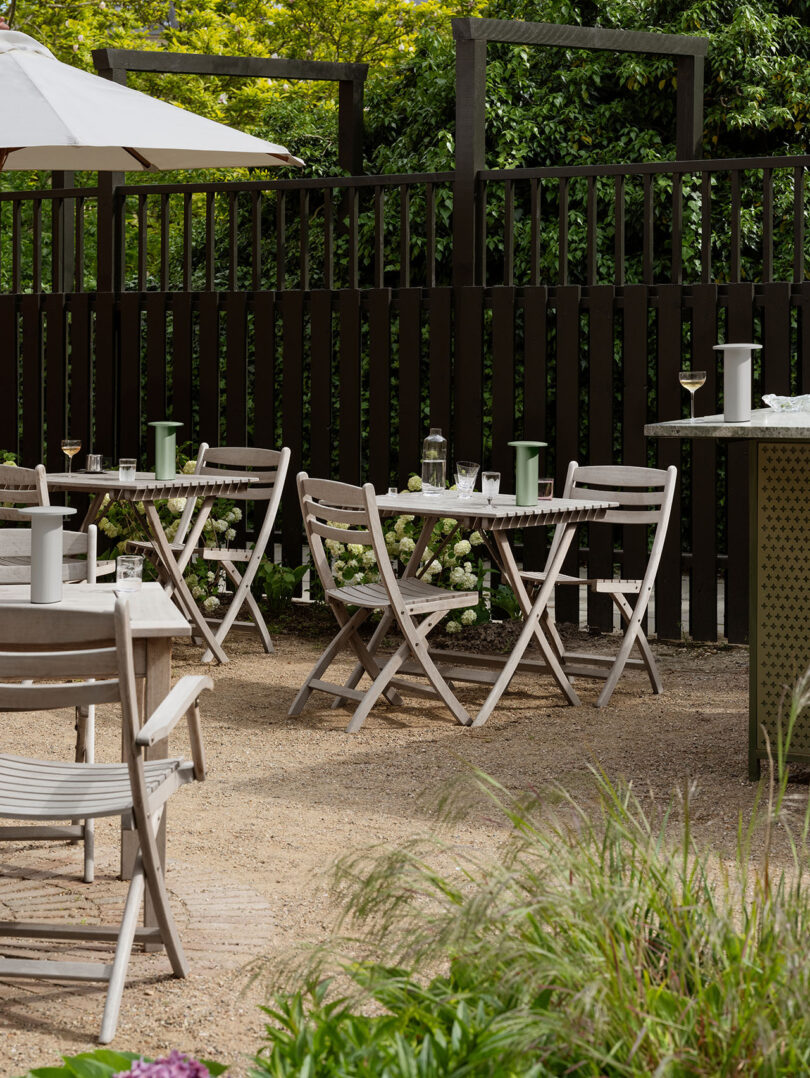 El patio al aire libre cuenta con mesas y sillas de madera de Cecile Manz con vasos y bebidas en el piso de grava, rodeado de vegetación y una cerca alta y oscura.