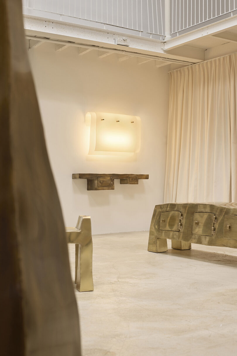 Minimalist room with light-colored walls, a sculptural brass bench and table, beige curtains, and a modern rectangular wall light fixture above a small shelf.