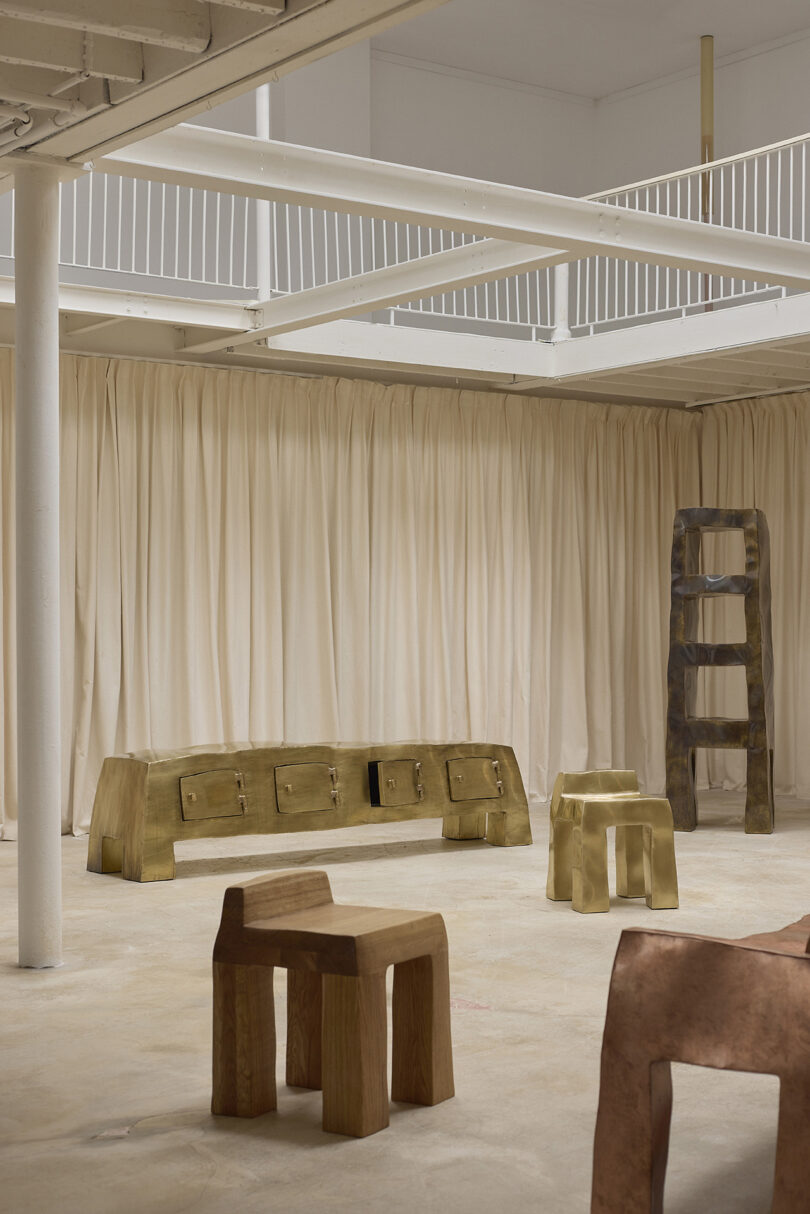 A minimalist interior features sculptural wooden furniture, including benches, chairs, stools, and a shelving unit, set against beige curtains in a spacious room with an upper balcony.
