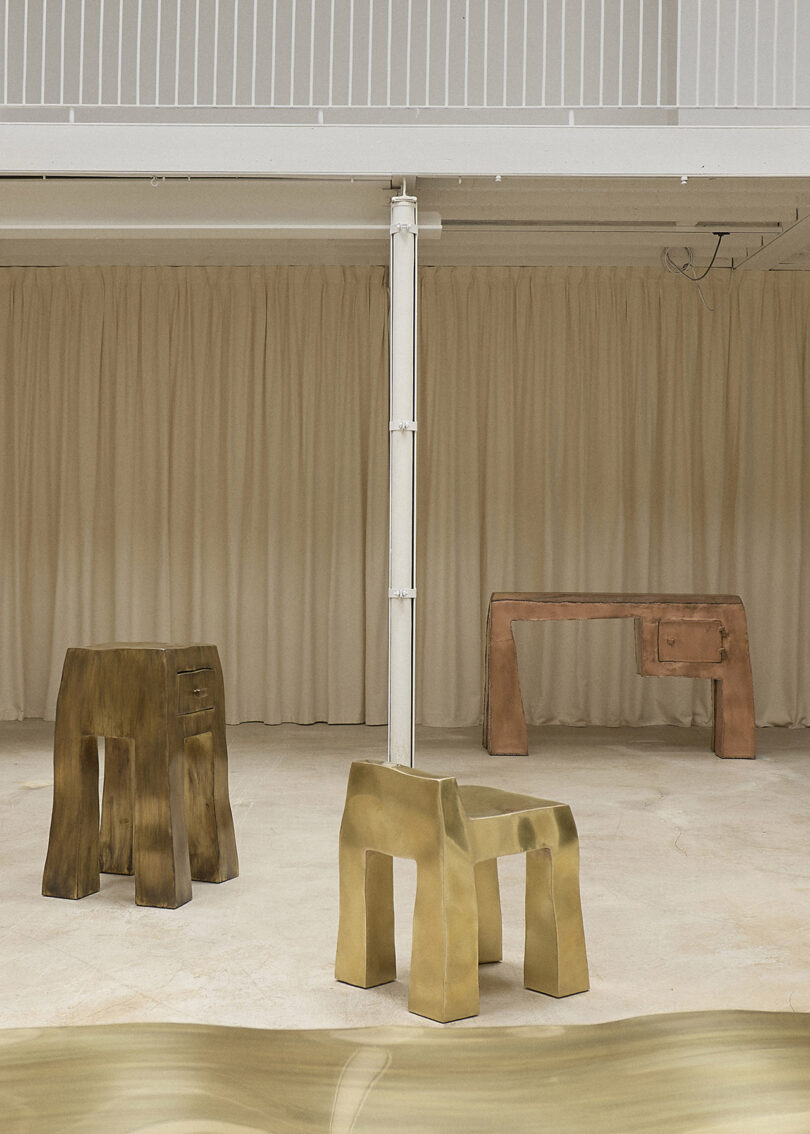 Three abstract, blocky tables in gold, brown, and copper tones are arranged in a minimal, beige room with a curtain backdrop and exposed white ceiling beams.