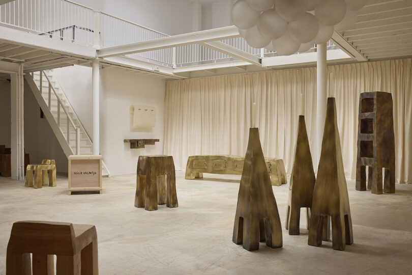 Spacious gallery with beige floors, cream curtains, and abstract wooden sculptures and furniture pieces arranged throughout the room. White staircase and railings visible in the background.