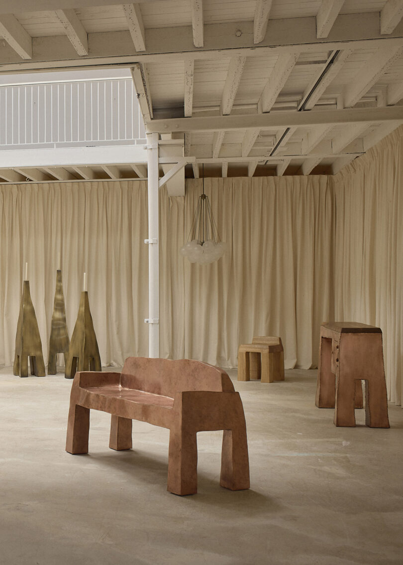 A minimal, beige-toned interior features sculptural furniture, draped curtains, tall candlesticks, and exposed ceiling beams under soft lighting.