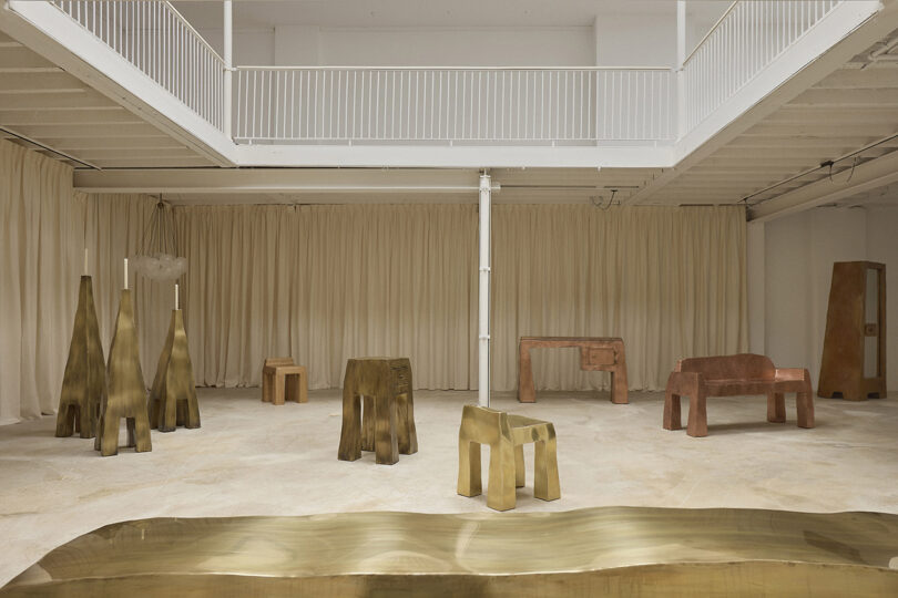 A spacious room with minimalist, sculptural furniture in metallic and earthy tones, set against beige curtains and a white industrial interior.